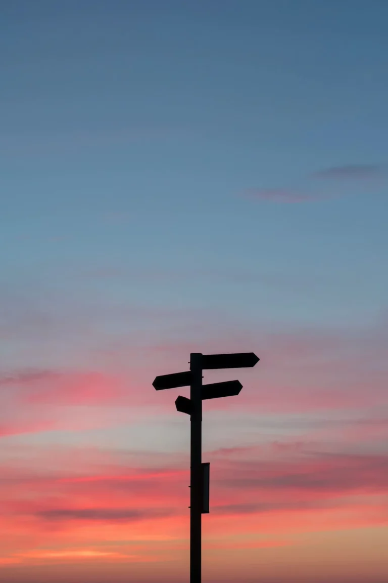 Un panneau de direction silhouetté contre un ciel au coucher du soleil aux couleurs bleu et rose.
