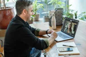 Un homme participe à une visioconférence avec une femme via son ordinateur portable, assis à une table en bois dans un environnement lumineux et végétalisé.