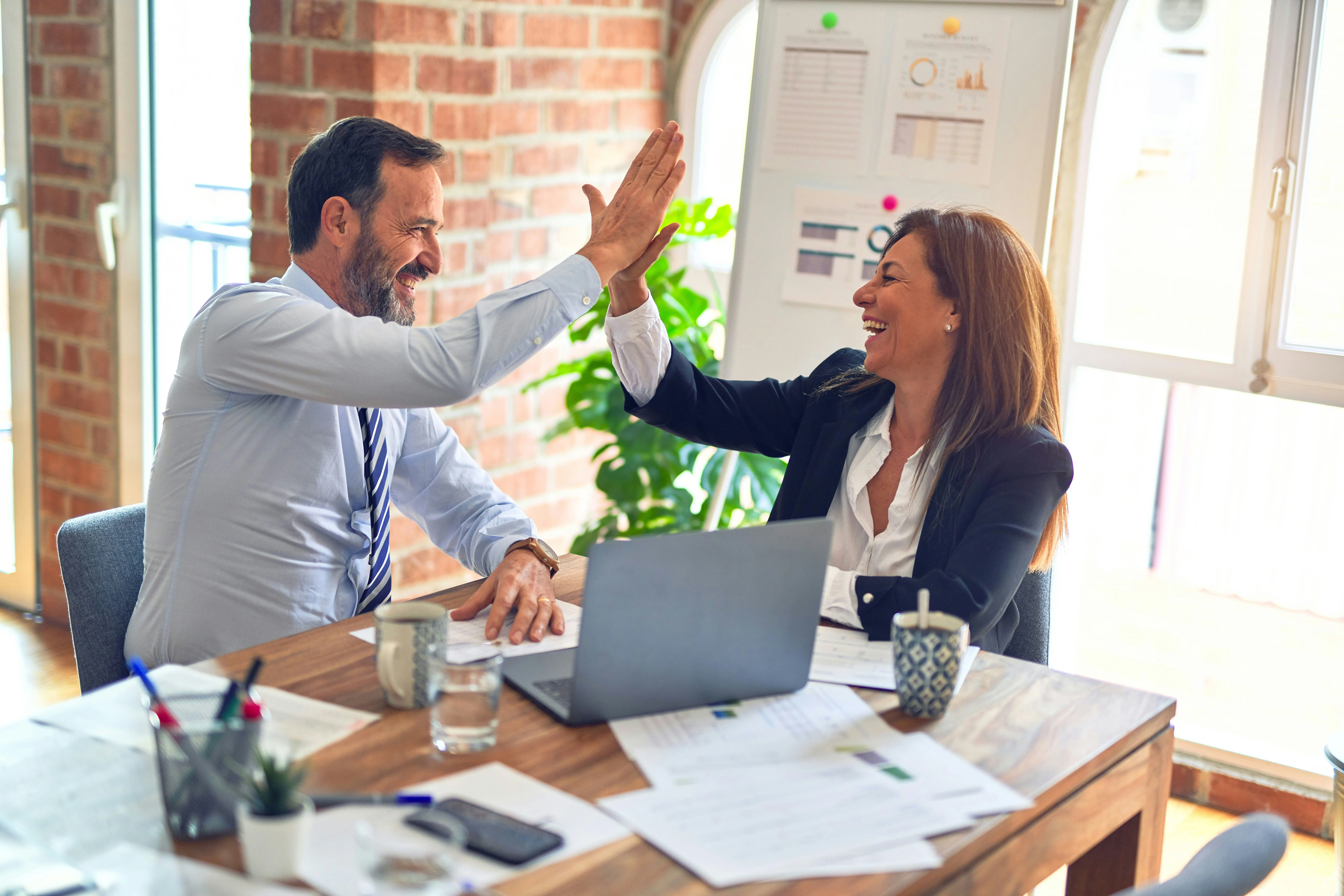 Deux collègues souriants se faisant un high-five autour d'une table de travail avec des ordinateurs portables et des documents, dans un bureau lumineux avec des plantes et un mur en brique.
